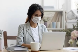 female employee wear medical protective mask from covid-19 coronavirus pandemic work on laptop in office