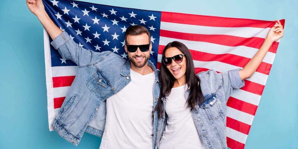 portrait of cheerful married people in eyewear eyeglasses holding us flag screaming wearing denim jeans jacket isolated over blue background
